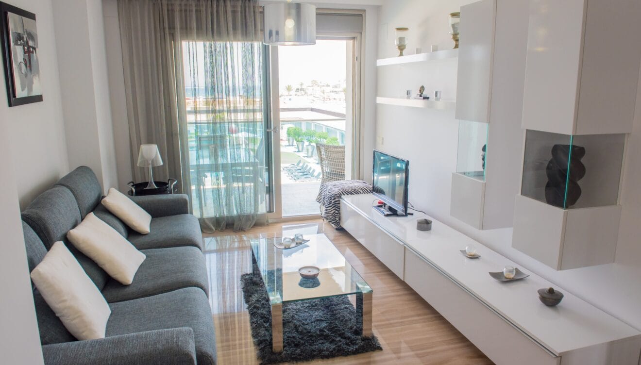 Salón moderno con sofá seccional gris y mesa de centro de cristal. Puertas correderas de vidrio dan a una terraza con vistas a la piscina y al mar. Suelos de mármol pulido