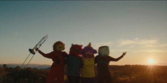Imagen: Cuatro personas con disfraces de monstruos de colores en un paisaje natural al atardecer, una de ellas levanta un trombón.