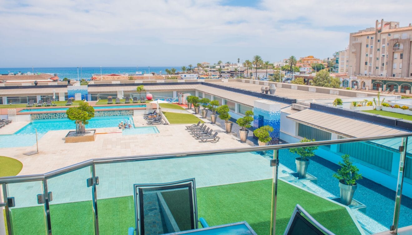 Vista elevada desde la terraza con barandilla de cristal, mostrando la piscina comunitaria, zonas de césped artificial y el mar Mediterráneo a lo lejos.