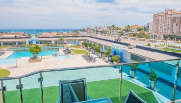 Vista elevada desde la terraza con barandilla de cristal, mostrando la piscina comunitaria, zonas de césped artificial y el mar Mediterráneo a lo lejos.