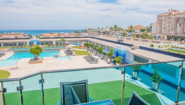 Imagen: Vista elevada desde la terraza con barandilla de cristal, mostrando la piscina comunitaria, zonas de césped artificial y el mar Mediterráneo a lo lejos.