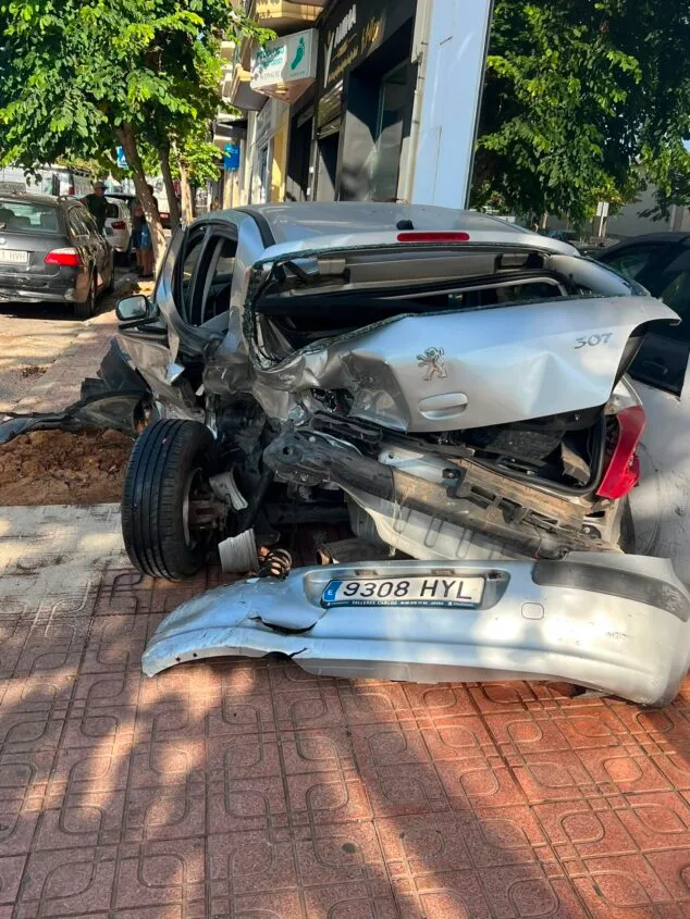 Imagen: Vista trasera de un coche Peugeot 307 plateado con graves daños estructurales y el parachoques desprendido sobre la acera tras un accidente.