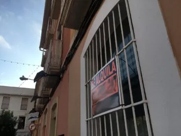 Fotografía de la fachada de un edificio con un gran cartel naranja y negro de 'SE ALQUILA' colgado en las rejas de una ventana o balcón.