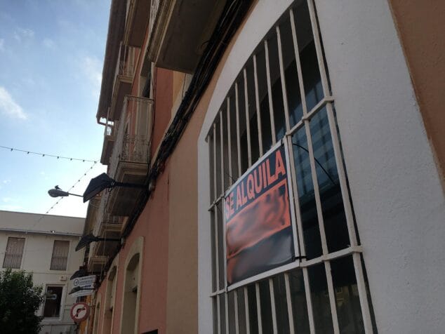Fotografía de la fachada de un edificio con un gran cartel naranja y negro de 'SE ALQUILA' colgado en las rejas de una ventana o balcón.