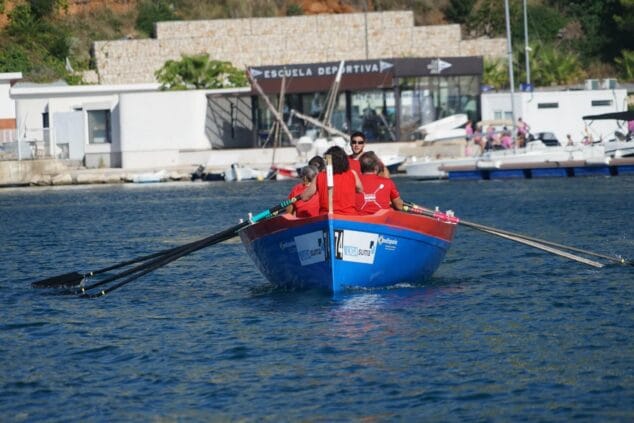 Imagen: Embarcación de remo azul con remeros de rojo de la Escuela Remo Valencia y logos de LIGA SUMA, remando en el puerto de Jávea.