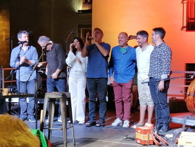 Imagen: Pep Botifarra cantando en un escenario al aire libre de noche acompañado por músicos.