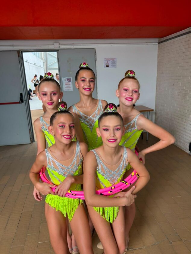 Imagen: Cinco gimnastas juveniles con maillots amarillo neón y tiaras posan juntas con un aro rosa.