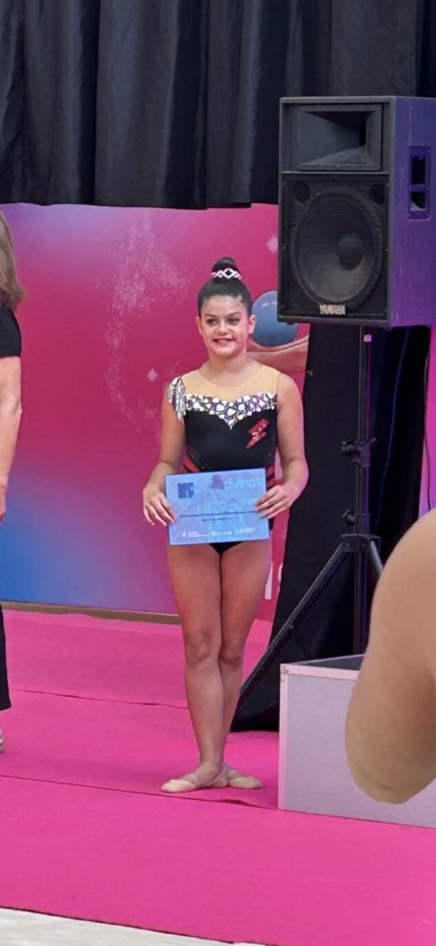 Imagen: La gimnasta alevín Fabiana de pie en un podio rosa sosteniendo un diploma azul tras su competición.