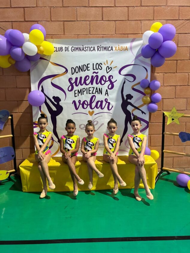 Imagen: Cinco jóvenes gimnastas del Club de Gimnàstica Rítmica Xàbia con maillots amarillos posan sentadas en un banco frente a un cartel del club.